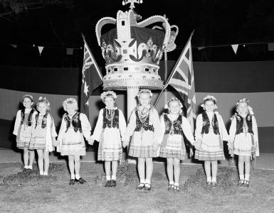 Children dressed in their national costume stand before a large model of the crown. 