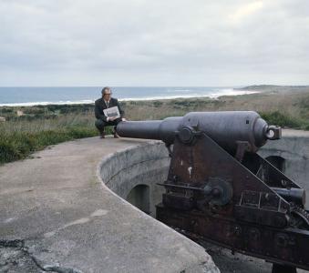 A man crouching beside a large fortified gun facing a coastline.