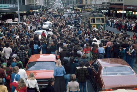 Cars and a tram surrounded by a crowd of people.