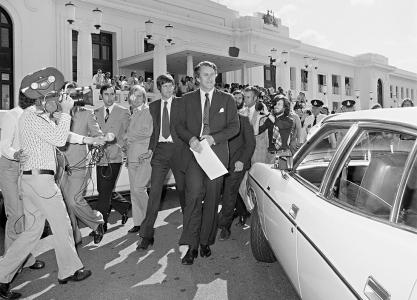 Malcolm Fraser walking toward a car at Parliament House followed by journalists and film crew.