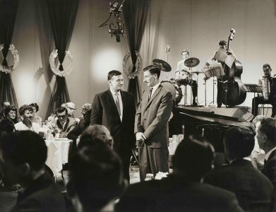 2 men talk amongst dinner tables beside a band on-stage