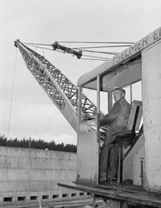 British migrant Albert Shaw in the cabin of the mobile crane he drives at Western Creek sewerage project. This crane lifted 10,000 cubic yards of concrete for Guthega Dam, in the Snowy Mountains Hydro Electric Scheme