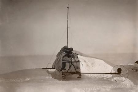 A sled in snow with mast and trailing wheel.