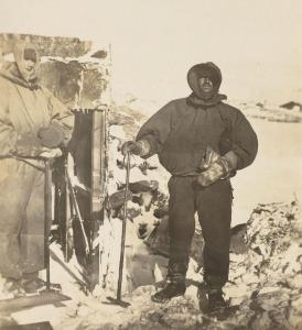 A man in polar clothing stands beside a timber hut.