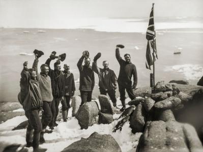 7 men standing beside the Union Jack flag on a snowy rocky mountain waving their beanies.