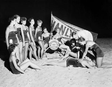 Surf lifesaving women receiving resuscitation training. Large Manly pennant behinf them. 