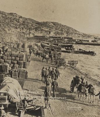 Anzac Beach, showing supplies on the left stacked in barrels, timber crates, and hessian bags. Mule drawn wagons moving along the beach.