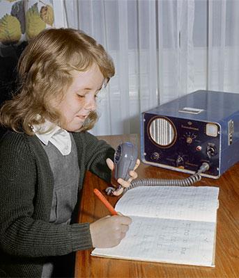 A young student writing in her study book while holding the microphone attached to a small radio tranceiver.