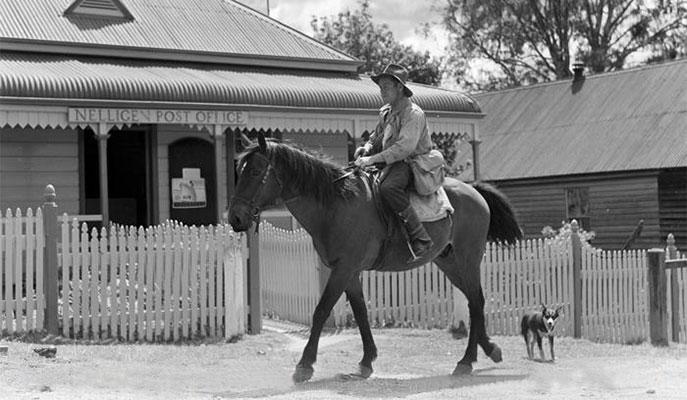 A postman riding a horse being followed by a dog