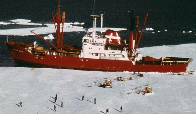 2 yellow helicopters and a group of expeditioners standing on ice beside red and white ship Nella Dan.