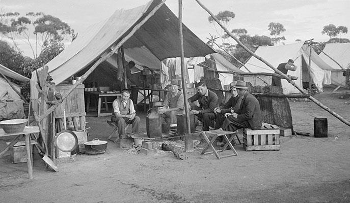 5 men sitting around a fire heating tin buckets in front of an open mess tent.