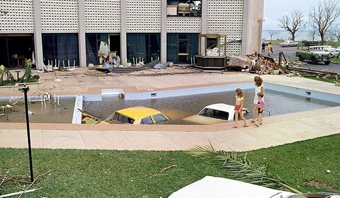 A woman and 2 children walk by 2 submerged cars in a multistorey hotel pool.