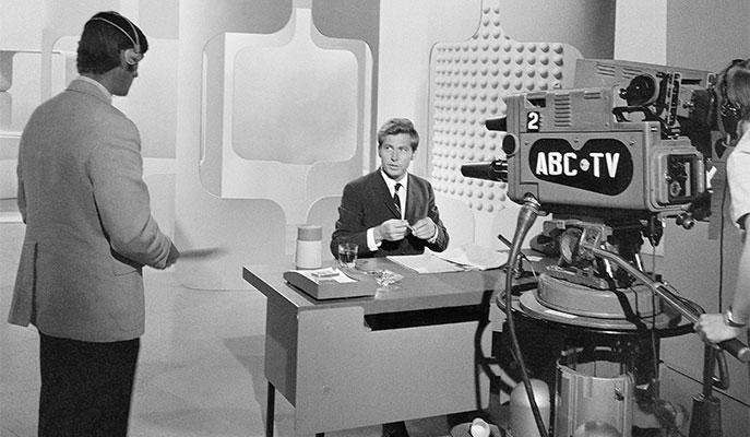 A man sitting at a desk infront of an ABC-TV camera.