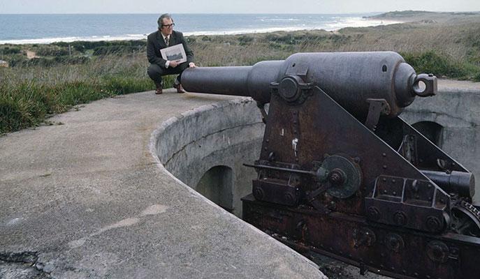 A man crouching beside a large fortified gun facing a coastline.