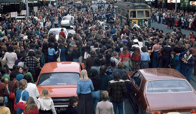 People crowding a street amongst cars and the 96 tram